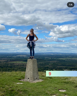 A white female with long brown hair smiles at the camera standing on a hill summit cairn. 